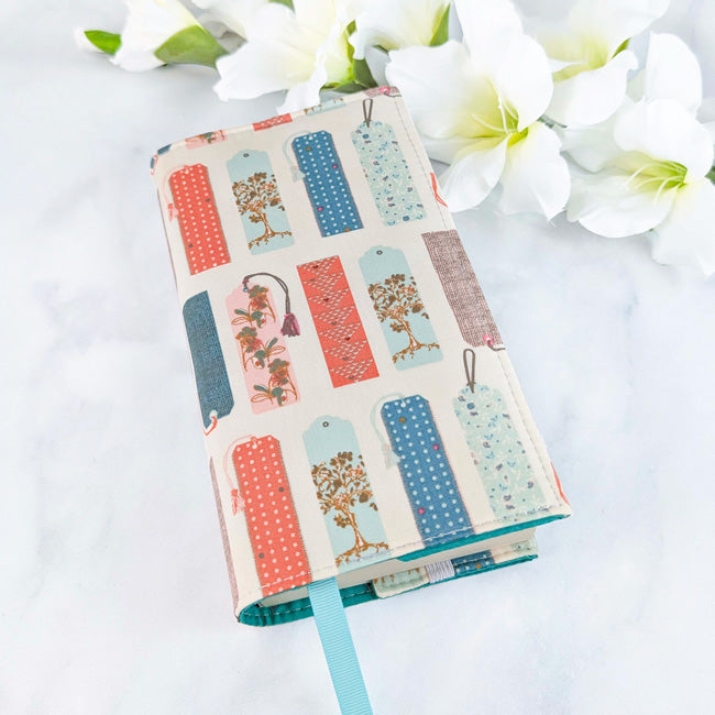 Fabric book cover featuring a pattern of colorful vintage-style bookmarks on a cream background, fitted on a book with a light blue ribbon bookmark, displayed beside white flowers.