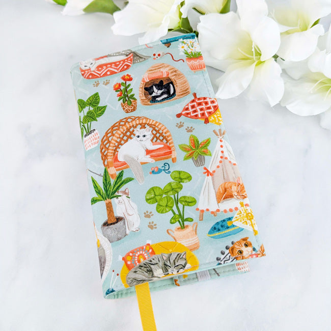 Fabric book cover featuring cozy illustrated cats lounging in hammocks, beds, and tents surrounded by houseplants, against a soft blue background.