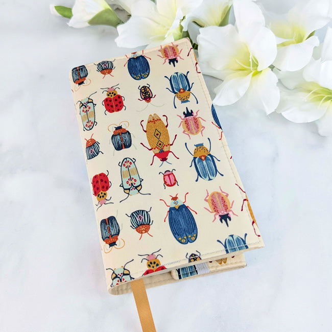 Fabric book cover with a whimsical pattern of colorful illustrated beetles and bugs on a cream background, shown with a tan ribbon bookmark and white flowers in the background.