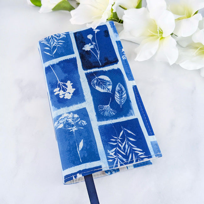 Fabric book cover with a blue and white botanical print resembling cyanotype art, featuring various leaves and flowers in rectangular frames, styled with white flowers.