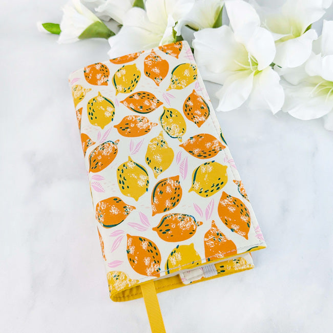 Fabric book cover with a cream background featuring a vibrant lemon and orange fruit pattern, fitted on a book with a yellow ribbon bookmark, displayed beside white flowers.