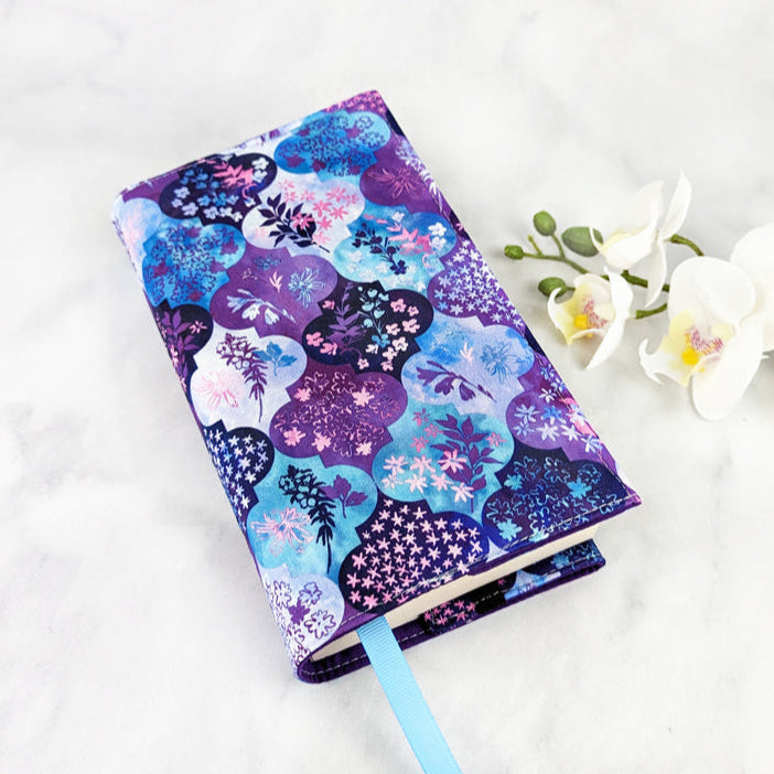 Fabric book cover with a purple, blue, and pink floral medallion pattern, fitted on a book with a light blue ribbon bookmark, displayed beside white flowers.