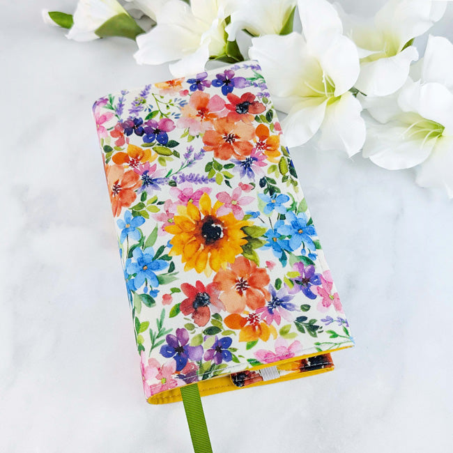 Fabric book cover featuring a vibrant floral pattern with sunflowers, lavender, and assorted wildflowers in bold, cheerful colors, shown with a green ribbon bookmark and white flowers in the background.