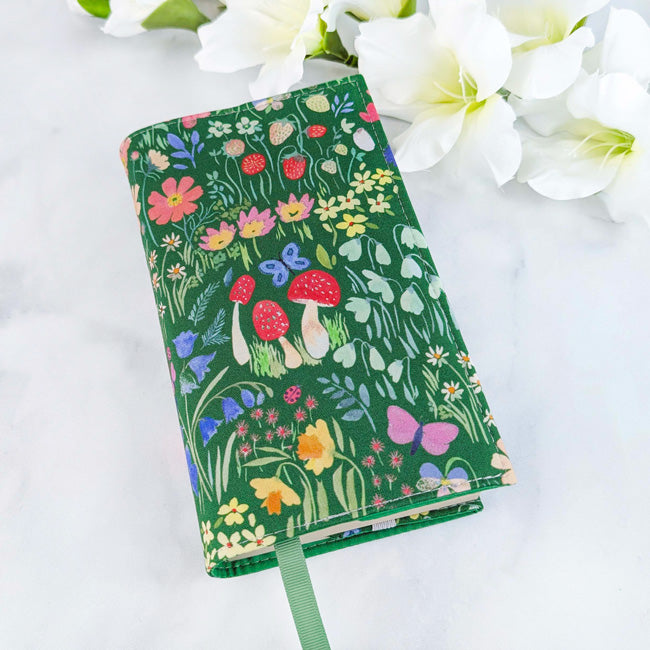 Fabric book cover with a green background featuring a colorful wildflower and strawberry pattern, fitted on a book with a green ribbon bookmark, displayed beside white flowers.