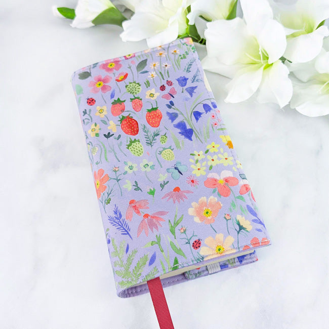 Fabric book cover with a purple background featuring strawberries, flowers, and greenery, fitted on a book with a red ribbon bookmark, displayed beside white flowers.
