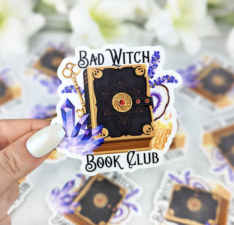 Vinyl sticker with the text “Bad Witch Book Club” featuring an ornate black spellbook with gold details, surrounded by crystals, lavender, a golden key, and candles, held by a hand with iridescent white nail polish.