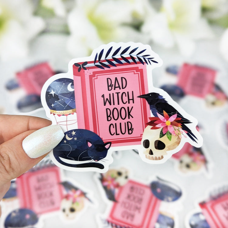 Vinyl sticker with the text “Bad Witch Book Club” on a pink book, decorated with a black cat, crow on a skull with flowers, and a crystal ball, held by a hand with iridescent white nail polish.