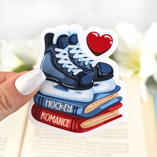 Vinyl sticker featuring blue ice skates on stacked books labeled “Hockey” and “Romance,” with a red heart above, held in hand over an open book.