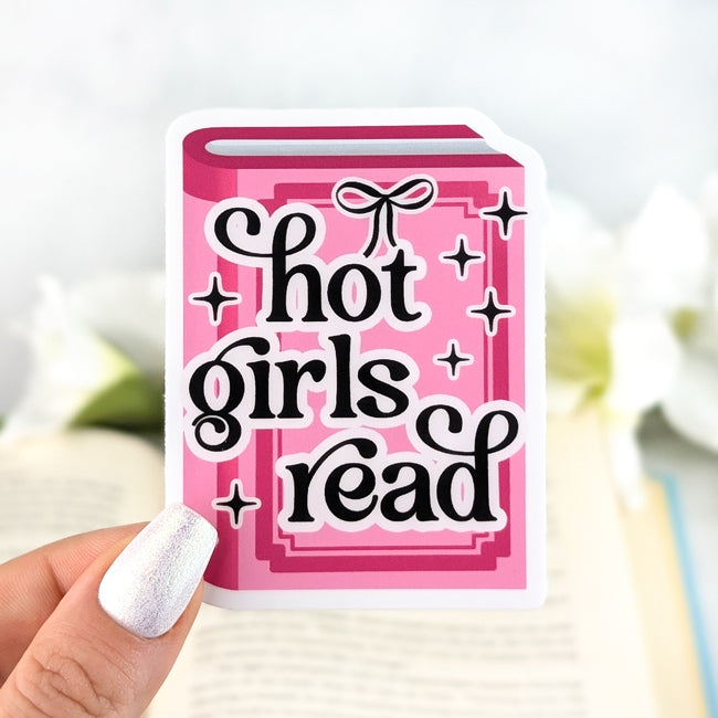 Pink book-shaped vinyl sticker with bold text “Hot Girls Read,” held in hand over an open book with white flowers in the background.