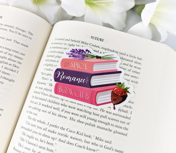 Spicy Romance Book Club vinyl sticker featuring illustrated stacked books, lavender, and a chocolate strawberry, placed on the pages of a novel.