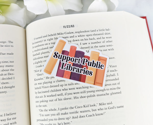 Support Public Libraries vinyl sticker featuring a row of illustrated books in warm colors, placed on the pages of a novel.