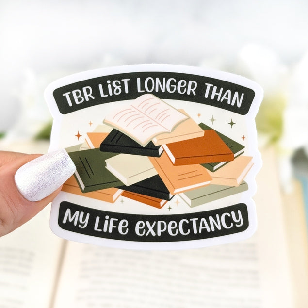 Vinyl sticker with stacked books and text “TBR List Longer Than My Life Expectancy,” held in hand over an open book background.