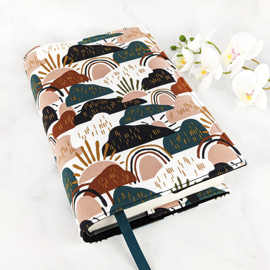 Fabric book cover with a modern abstract design featuring overlapping hills, suns, and rainbows in earthy tones of rust, olive, blush, and black on a white background, finished with a dark green ribbon bookmark.