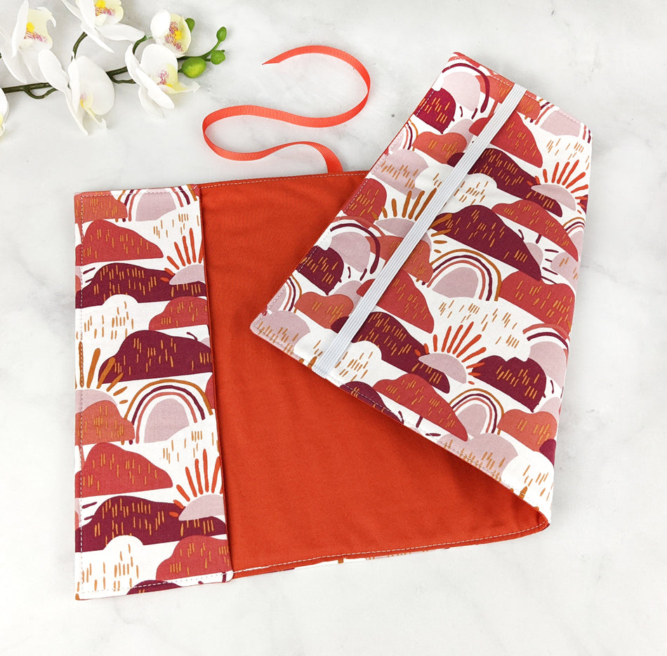 Fabric book cover with a whimsical abstract design of suns, rainbows, and hills in warm shades of terracotta, blush, and maroon on a white background, finished with an orange ribbon bookmark.