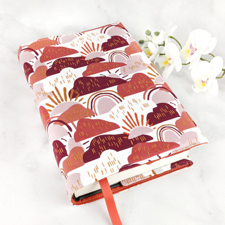 Fabric book cover with a whimsical abstract design of suns, rainbows, and hills in warm shades of terracotta, blush, and maroon on a white background, finished with an orange ribbon bookmark.