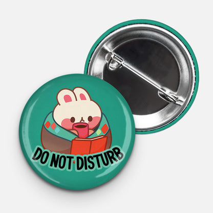 Round teal pinback button featuring a cute cartoon bunny wrapped in a blanket, holding a mug and reading a book. Bold black text at the bottom reads “DO NOT DISTURB.” The back of the button with a metal pin is also visible.