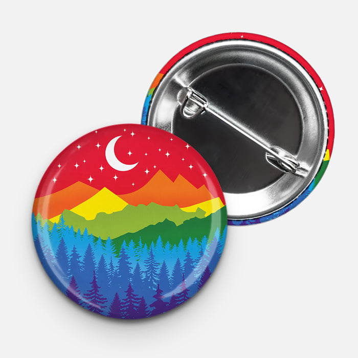 Round 1.75" pinback button with a colorful mountain and forest scene in the rainbow pride flag colors, featuring a crescent moon and star-filled sky. The pin’s backside with a metal clasp is partially shown beside it.
