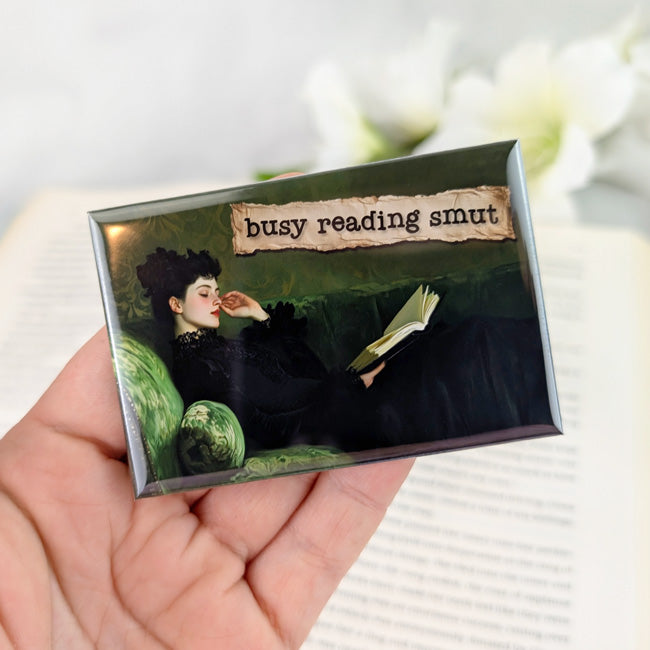 Rectangular magnet showing a vintage-style painting of a woman lounging on a green velvet sofa in a black dress, reading a book. A torn-paper label above her reads “busy reading smut.” The magnet is held in a hand with iridescent white nail polish, against a blurred open book background.