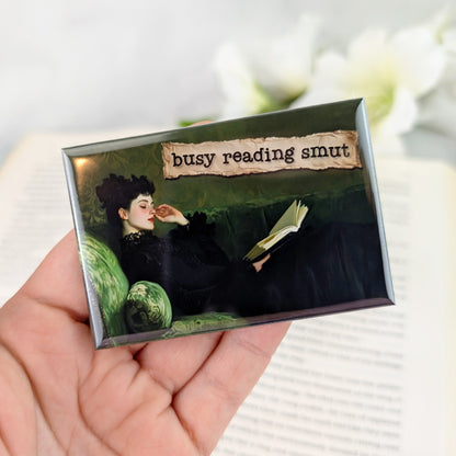 Rectangular magnet showing a vintage-style painting of a woman lounging on a green velvet sofa in a black dress, reading a book. A torn-paper label above her reads “busy reading smut.” The magnet is held in a hand with iridescent white nail polish, against a blurred open book background.