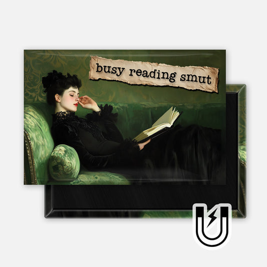 Rectangular magnet featuring a vintage-style painting of a woman reclining on a green velvet sofa while reading a book, with the text “busy reading smut” on a torn paper banner overlay. The woman wears a black dress, and the background is a muted green patterned wall.