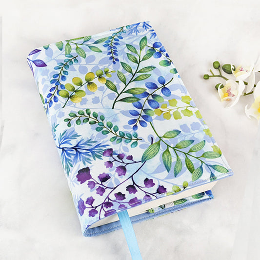 Fabric book cover featuring a delicate watercolor-style botanical print with leaves and branches in shades of green, blue, yellow, and purple on a soft sky-blue background, finished with a light blue ribbon bookmark.