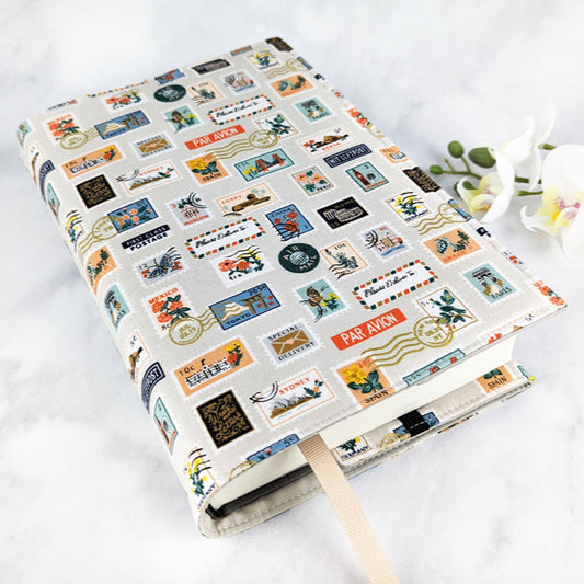 Fabric book cover featuring a travel-themed print of colorful vintage-style postage stamps and travel labels on a light gray background, with a beige ribbon bookmark.