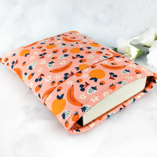 medium padded book sleeve with pocket. fabric showcases oranges, bananas, strawberries, blueberries and flowers on a light pink background.