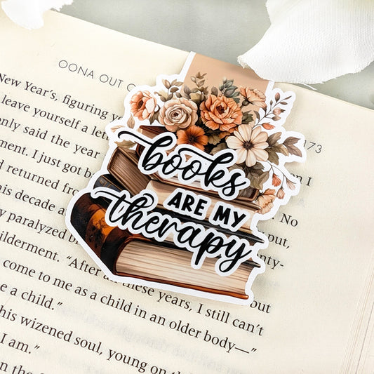 Magnetic bookmark with 'Books Are My Therapy' and floral design on a stack of books, placed on an open book.