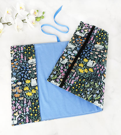 Fabric book cover featuring a whimsical print of white cats lounging and playing among colorful wildflowers on a black background, finished with a blue ribbon bookmark.