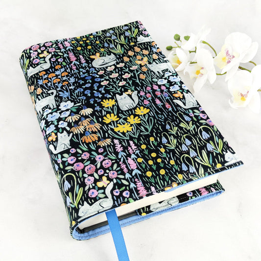 Fabric book cover featuring a whimsical print of white cats lounging and playing among colorful wildflowers on a black background, finished with a blue ribbon bookmark.