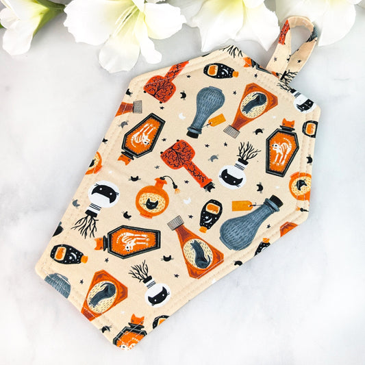Coffin-shaped quilted potholder with loop handle, featuring a whimsical pattern of apothecary bottles and specimen jars—bats, bones, cats, herbs—on a warm tan background. Displayed on a marble surface beside white flowers.