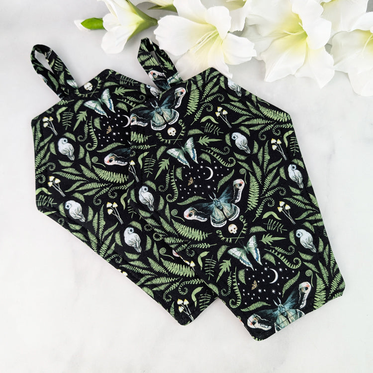 Two Coffin-shaped quilted potholders with loop handle, displayed on a marble surface beside white flowers. The fabric shows a dark botanical and moth pattern, and the potholder is positioned diagonally for a clear, top-down view.