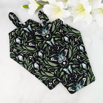 Two Coffin-shaped quilted potholders with loop handle, displayed on a marble surface beside white flowers. The fabric shows a dark botanical and moth pattern, and the potholder is positioned diagonally for a clear, top-down view.