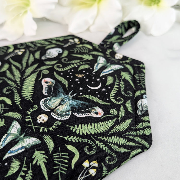 Coffin-shaped quilted potholder with loop handle, displayed on a marble surface beside white flowers. The fabric shows a dark botanical and moth pattern, and the potholder is positioned diagonally for a clear, top-down view.