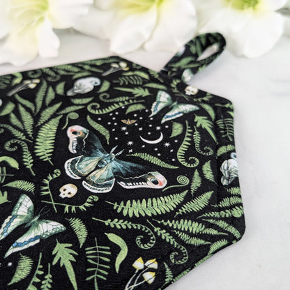Coffin-shaped quilted potholder with loop handle, displayed on a marble surface beside white flowers. The fabric shows a dark botanical and moth pattern, and the potholder is positioned diagonally for a clear, top-down view.