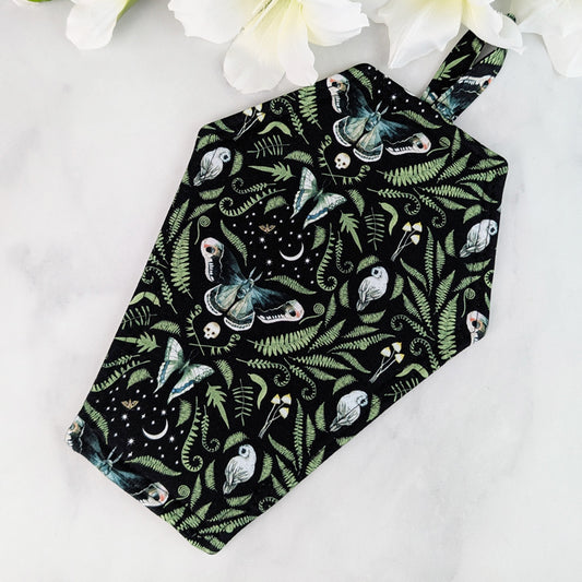 Coffin-shaped quilted potholder with loop handle, displayed on a marble surface beside white flowers. The fabric shows a dark botanical and moth pattern, and the potholder is positioned diagonally for a clear, top-down view.