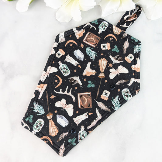 Coffin-shaped quilted potholder with loop handle, featuring a black fabric pattern of witchy curiosities—brooms, crystals, skulls, potion bottles, antlers, and crescent moons—displayed on a marble surface beside white flowers.