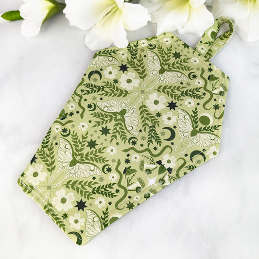 Coffin-shaped quilted potholder with loop handle, featuring a green botanical and celestial pattern of moths, stars, moons, and leaves. Displayed on a marble surface beside white flowers.