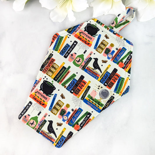 Coffin-shaped quilted potholder with loop handle, featuring a colorful pattern of potion bottles, spell books, cauldrons, ravens, and apothecary labels on a cream background. Displayed on a marble surface beside white flowers.