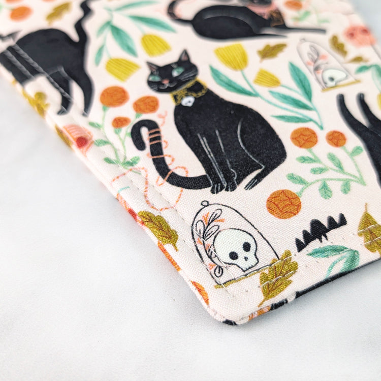 Coffin-shaped quilted potholder with loop handle, featuring a whimsical pattern of black cats, yarn balls, leaves, and skull motifs on a pale background. Displayed on a marble surface beside white flowers.