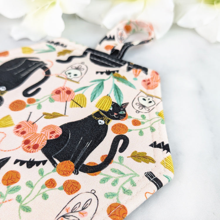 Coffin-shaped quilted potholder with loop handle, featuring a whimsical pattern of black cats, yarn balls, leaves, and skull motifs on a pale background. Displayed on a marble surface beside white flowers.
