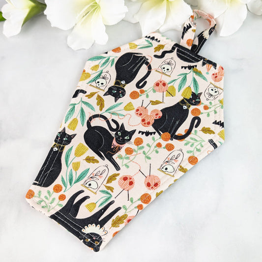 Coffin-shaped quilted potholder with loop handle, featuring a whimsical pattern of black cats, yarn balls, leaves, and skull motifs on a pale background. Displayed on a marble surface beside white flowers.