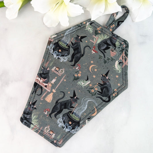 Coffin-shaped quilted potholder with loop handle, featuring a gray fabric pattern of black cats wearing witch hats, stirring cauldrons, and surrounded by potions, frogs, and crescent moons. Displayed on a marble surface beside white flowers.