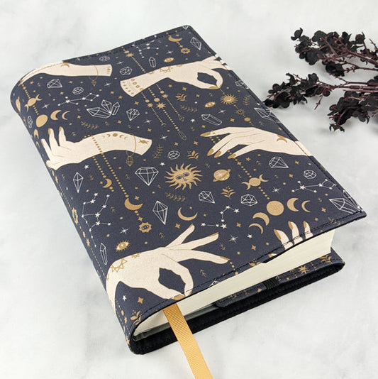 Fabric book cover with a celestial design featuring cream-colored hands, golden suns, moons, stars, constellations, and crystals on a dark navy background. 