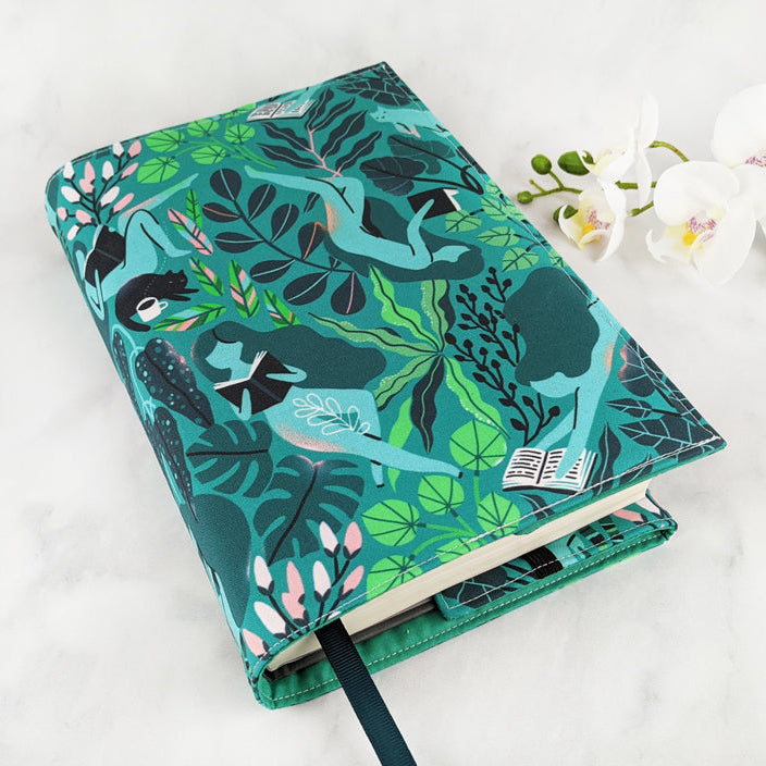 Fabric book cover with a vibrant botanical design featuring illustrated women reading books while lounging among lush green foliage and soft pink flowers, finished with a dark teal ribbon bookmark.