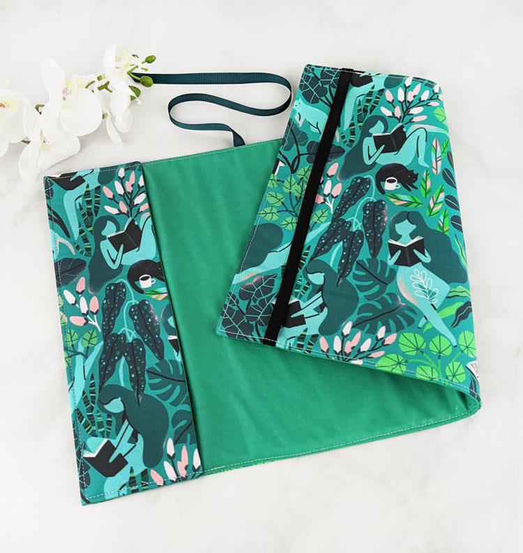 Fabric book cover with a vibrant botanical design featuring illustrated women reading books while lounging among lush green foliage and soft pink flowers, finished with a dark teal ribbon bookmark.