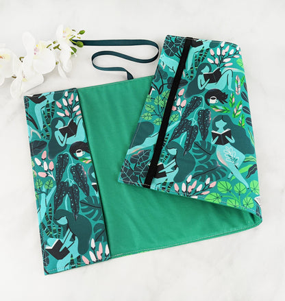Fabric book cover with a vibrant botanical design featuring illustrated women reading books while lounging among lush green foliage and soft pink flowers, finished with a dark teal ribbon bookmark.