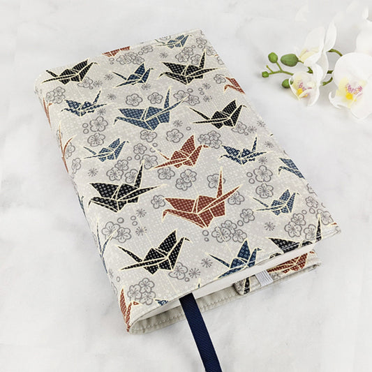 Fabric book cover featuring a traditional Japanese-inspired design with patterned origami cranes in navy, black, and rust tones, surrounded by plum blossoms on a soft beige background, finished with a dark blue ribbon bookmark.
