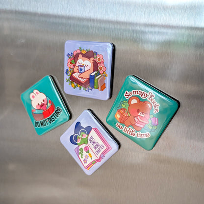  Four colorful square magnets with cute animal designs on a metallic surface. The magnets feature: a bunny reading with "DO NOT DISTURB," an owl reading a book surrounded by flowers, a bear carrying books with "So many books, so little time," and a frog in a witch hat with "JUST ONE MORE CHAPTER."