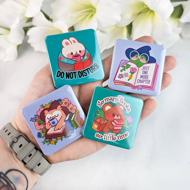 A hand holding four colorful square magnets with cute animal designs. The magnets feature: a bunny reading with "DO NOT DISTURB," a frog in a witch hat with "JUST ONE MORE CHAPTER," an owl reading a book surrounded by flowers, and a bear carrying books with "So many books, so little time."